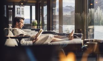 A man is sitting relaxed in a bathrobe on a couch and reading a book. The windows offer a view of the landscape outside.