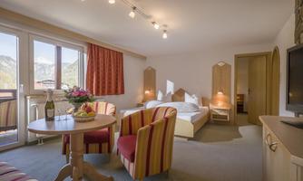 A cozy hotel room with a double bed, a small table, and two striped chairs. Large windows offer a view of the mountains.