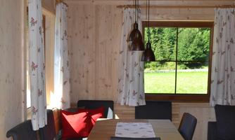 A cozy dining area in a wooden cabin with a large table and comfortable chairs. Large windows offer a view of the green landscape outside.