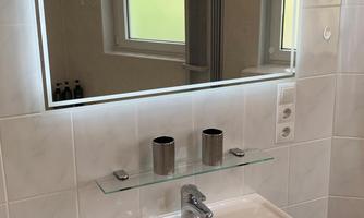 A modern bathroom with a sink and a lit mirror. Next to it is a small towel holder and some bathroom utensils are neatly arranged.