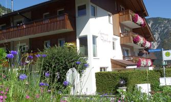 A beautiful building with wooden cladding and balconies, blooming plants, and a well-maintained garden. The surroundings are enclosed by mountains, creating a peaceful atmosphere.