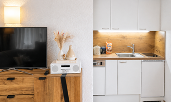 A modern living room with a wooden dresser and a television. The kitchenette is bright and functional, with white cabinets and a wooden countertop.