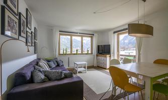 A modern living room with a comfortable sofa, a dining table, and large windows overlooking the mountains. There are pictures on the wall and plenty of daylight.