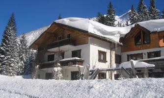 A snow-covered house in a winter landscape. Surrounded by tall, green pine trees and clear blue sky.