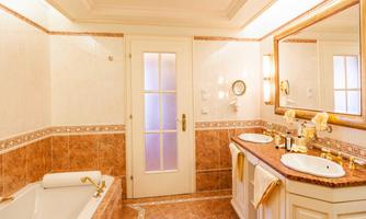 An elegant bathroom with marbled walls and a large mirror. There is a bathtub as well as two sinks with gold fixtures.