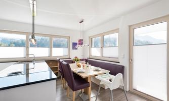 A modern kitchen and dining area with a large table and purple chairs. Large windows provide plenty of natural light and a view of the landscape.