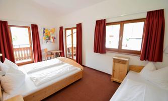 A bright room with two beds and red curtains. There are windows overlooking a balcony, as well as a small table and chairs.