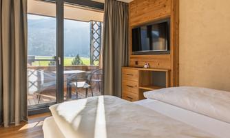 A modern room with a large window and a balcony that offers a beautiful view of the mountains. There is a cozy bed and a wooden dresser.