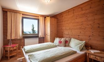 A cozy bedroom with wood paneling and a large window. The room is furnished with a double bed, pillows, and a small table.