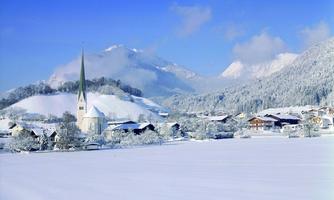 A snowy landscape with a small village and a church. In the background, high mountains can be seen under a clear blue sky.