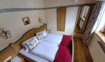 A cozy bedroom with a double bed, two pillows, and a red blanket. There is a mirror and daylight shining through the curtains.