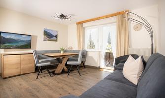 A cozy living space with a sofa, a dining area, and large windows. Pictures of landscapes hang on the wall, and the room is bright and inviting.