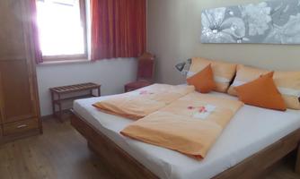A cozy bedroom with a double bed and orange pillows. The walls are light, and there is a large window with red curtains.
