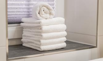 A stack of white towels is neatly arranged on a shelf in a bathroom. In the background, bright walls and a window with blinds can be seen.
