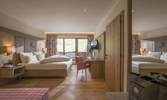 A modern hotel room with wooden furniture and light curtains. There is a large bed, a television, and a comfortable armchair.
