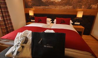 A cozy hotel room with a double bed and red pillows. In the foreground lies a towel and a bag with the print "DAS SPREICHL".
