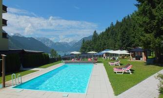 A swimming pool surrounded by green areas and loungers. In the background, mountains and a bright blue sky are visible.