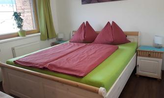 A cozy bedroom with a large bed and red pillows. Next to the bed is a nightstand and a window with green curtains.