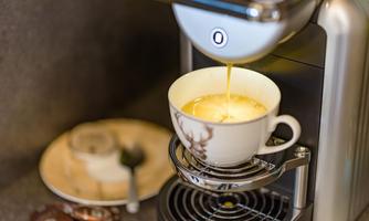 A coffee machine brews freshly brewed coffee into a decorative cup. Next to it lies a plate with a spoon and some coffee capsules.