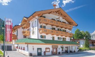 A charming hotel in alpine style with wooden balconies. Surrounded by beautiful trees and a blue sky.
