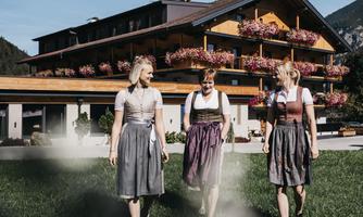 Three women in traditional dresses are walking across a meadow in front of a building with balcony flowers. The surroundings are mountainous and sunny.