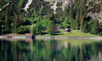 A picturesque landscape by the lake with several wooden houses and dense trees. The calm water reflects the surroundings.