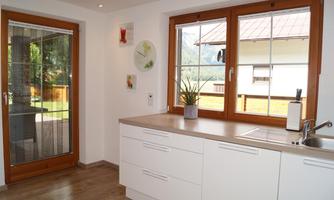 A modern kitchen with white cabinets and a countertop. Large windows offer a view of nature.