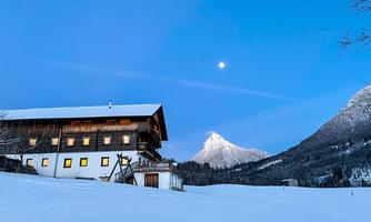 A picturesque mountain cabin in the snow, surrounded by majestic mountains. The clear sky and the moon give the scene a tranquil atmosphere.