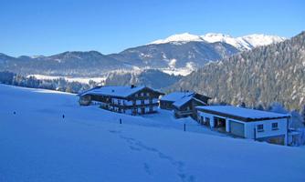 A snowy landscape with traditional wooden houses. In the background, majestic mountains and a clear blue sky can be seen.