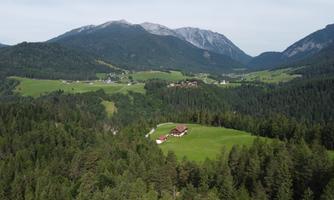 A picturesque landscape with green meadows and mountains in the background. On a hill stands a red farmhouse, surrounded by trees.