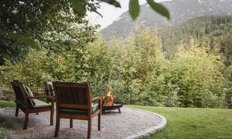 A cozy outdoor area with two chairs and a fire pit. In the background, wooded mountains and a green landscape can be seen.