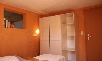 A simple, bright room with orange walls. It contains a bed, a table, and a white wardrobe.