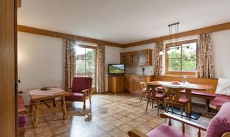 A cozy living room with wooden furniture and large windows. There is a dining table, comfortable chairs, and a television.