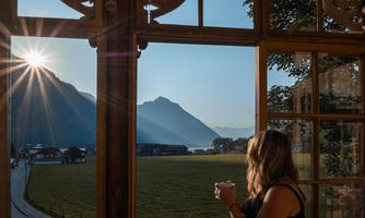 A woman looks out of a window at a picturesque landscape with mountains and the sun rising. Outside, green meadows and a clear sky can be seen.