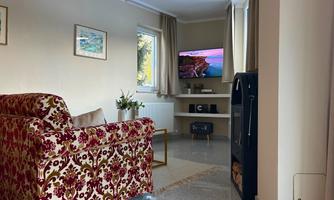 A cozy living room with a red patterned sofa and a TV on the wall. Next to the sofa is a small table and a plant on the window sill.