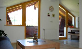 A bright living room with large windows and a cozy table. In the background, plants and another room are visible.