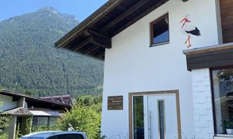 A modern building with a white car in front and a mountain landscape in the background. The facade features a stylized stork.