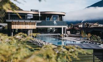 A modern building with a large pool and a view of the mountains. The surroundings are green and inviting.