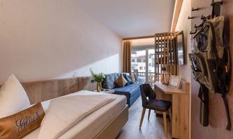 A cozy hotel room with a single bed and a seating area. The room has large windows with access to a balcony and modern furniture.
