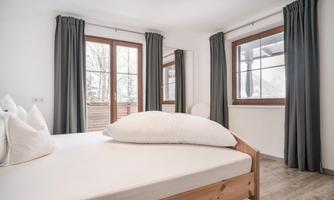 A bright bedroom with a large window and dark curtains. The bed has white bedding and is simply but stylishly furnished.