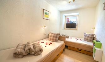 A cozy bedroom with two single beds and decorative pillows. Rose petals are scattered on the floor, and there is a window view outside.