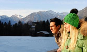 A young couple looks out at a snowy landscape with mountains in the background. They are wearing warm clothing and enjoying the view.
