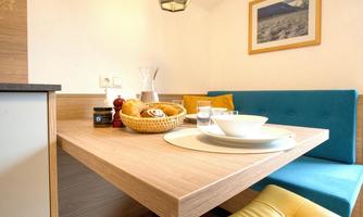 An inviting dining area with a bright wooden table and cozy seating. On the table are dishes, a basket of rolls, and beverages.