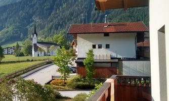 A picturesque landscape with mountains in the background and traditional houses. In the foreground, there is a balcony with garden fruits and trees.
