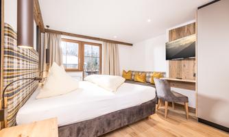 A modern bedroom with a large bed and a television. The room is bright and friendly with wooden details and cozy cushions.
