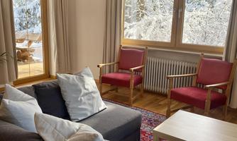 A cozy living area with a gray sofa and red chairs. Large windows provide a view of a snowy landscape.