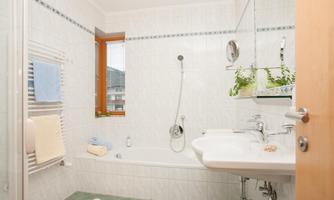 A modern bathroom with a bathtub and a shower. There are bright tiles and a small window opening looking outside.