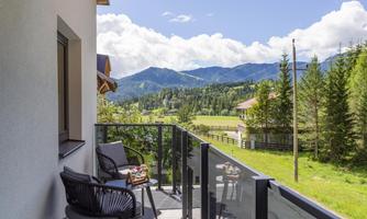 A balcony with two chairs and a table, surrounded by a beautiful mountain landscape. In the background, trees and mountains are visible under a blue sky.