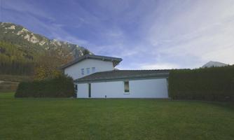A modern house surrounded by a well-kept garden. In the background, high mountains and a clear sky can be seen.