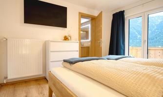 A modern bedroom with a large bed, a TV, and a wardrobe. The windows offer a view of the mountains and let in a lot of light.